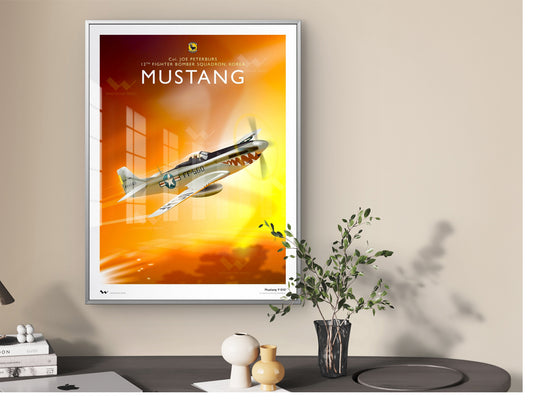 A framed art print featuring a vintage Mustang airplane against a warm orange and yellow sky, displayed on a beige wall above a dark table with decorative objects and a plant.
