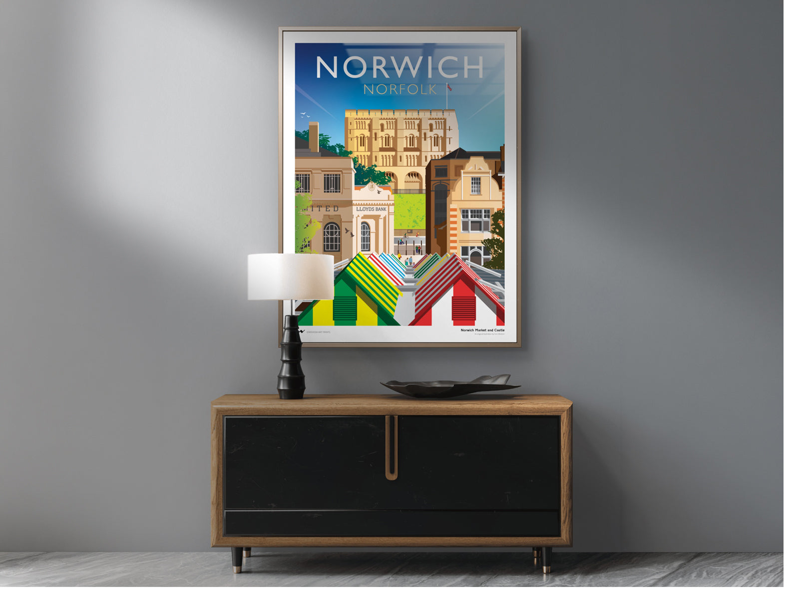 Framed art print of Norwich on a wall above a wooden cabinet with a lamp and decorative bowl.